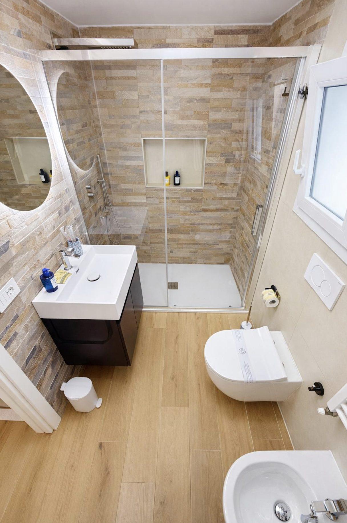 Designer bathroom with walk-in shower, natural stone tiles, oval mirror, and bidet in Mare apartment